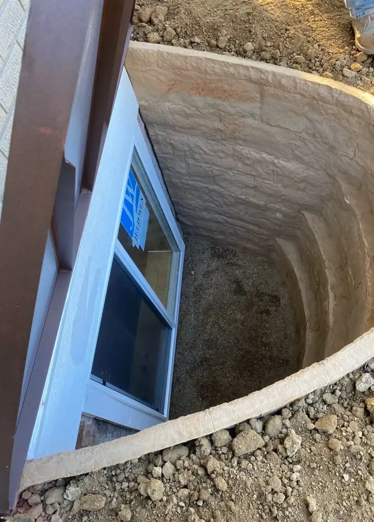 Egress Window Well Covers: cover lifted from egress well with half-oval, false stone walls and sand bottom, egress window in well placed into foundation below ground level